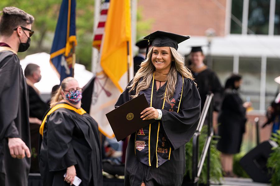 A female student walks and smiles with her diploma at the in-person recognition ceremony.
