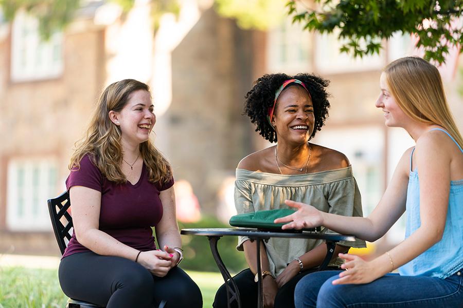 Three students sit at a table on campus and talk.