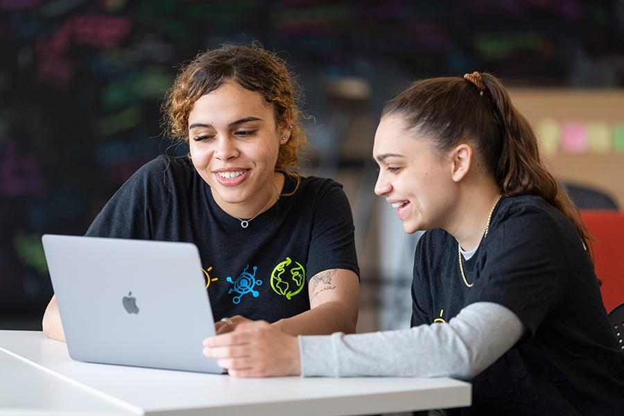 Two students work on a laptop in the Hynes Institute and smile.