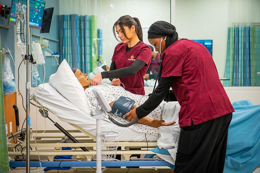 Two Iona nursing students practice reading vitals on a mannequin.