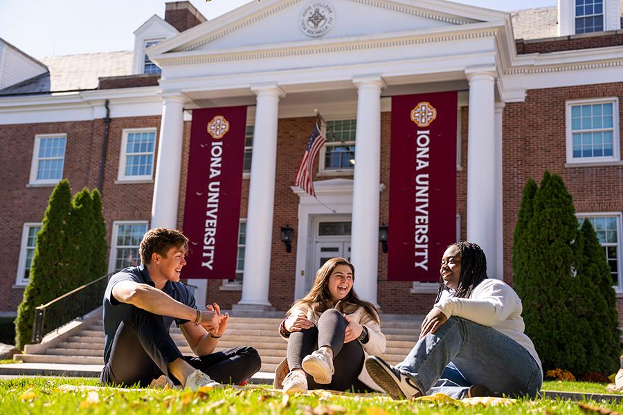 Three students sit in front of McSpedon Hall on a sunny day and smile and talk.