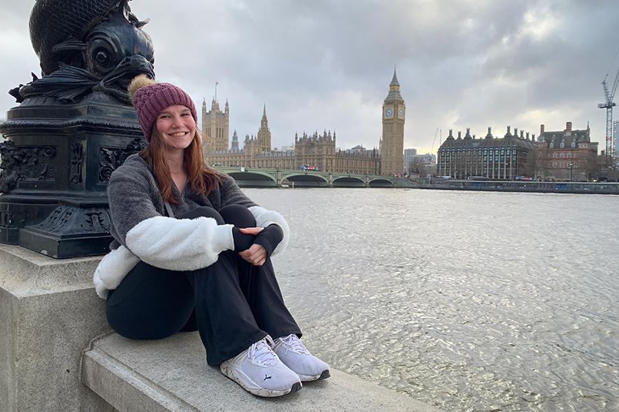 A student sits by the river with Big Ben in the background and smiles.