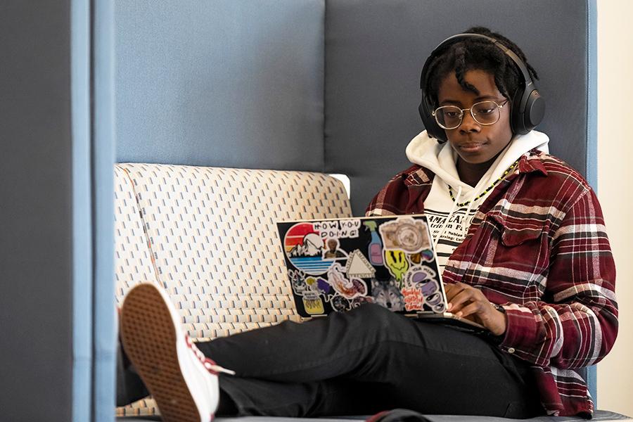 A student works on her laptop on a couch with headphones on.