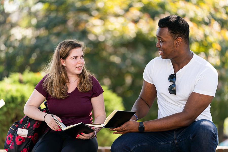 Two students sit outside and talk and smile on a sunny day.