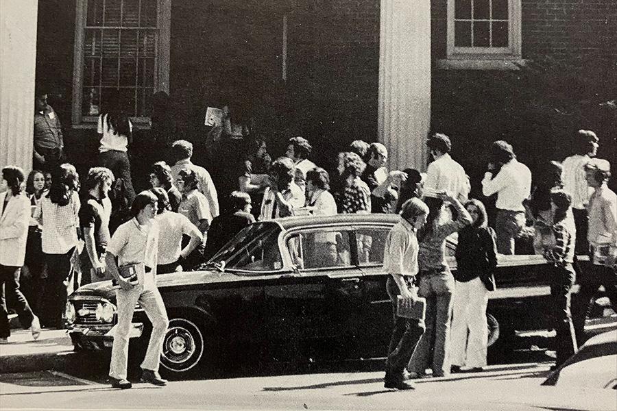 An old photo of Spellman Hall with students walking between classes and a car.
