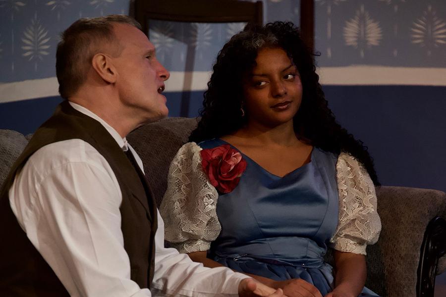 Two male actors sitting on the couch, the male actor is speaking while the female actor looks at the male actor, in the play "Creditors."