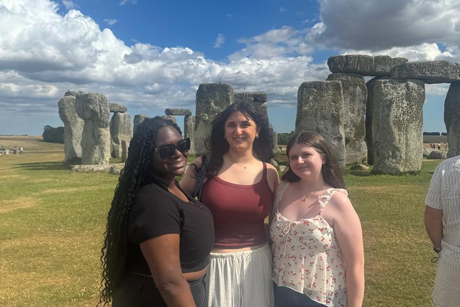 Three students smiling and posing for a group photo, in front of the historical landmark known as the Stonehenge in England.