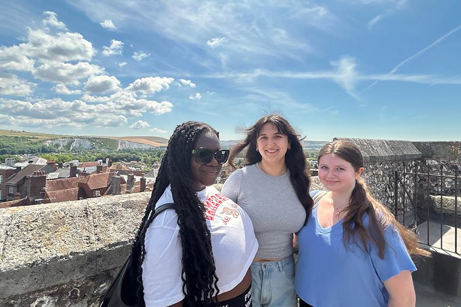 Three students smiling and posing for a group photo outside on a balcony, with the Lewes, England land in the background.