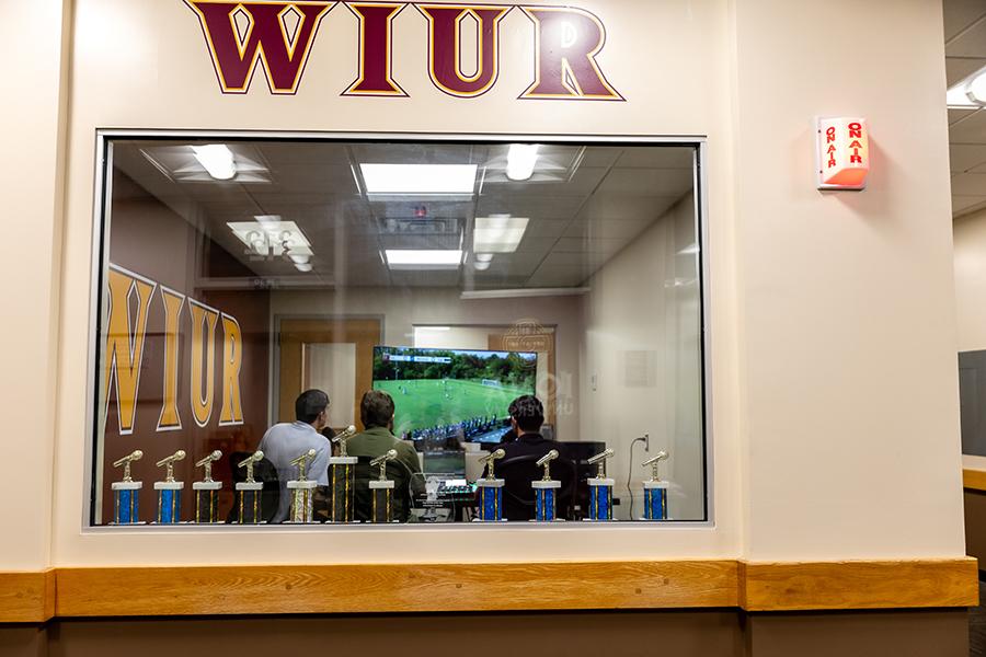 A photo of the Iona University Radio Station Room, looking through a glass window, where three male students are doing a live radio broadcast.