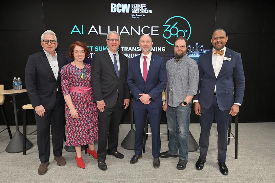 A group of six panelists and moderators stand together for a photo in front of the "AI Alliance 360" digital backdrop at the Business Council of Westchester summit.