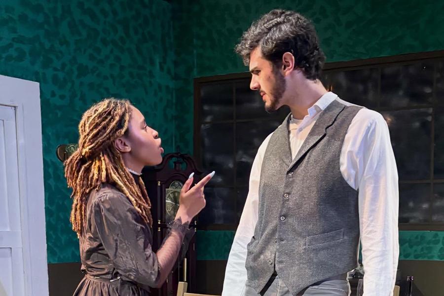A young woman in a dark dress points her finger accusingly at a tall man in a grey vest and white shirt in a tense scene from Henrik Ibsen's play "Ghosts."
