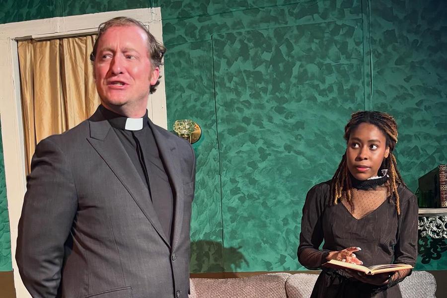 A man dressed as a priest speaks while a young woman holds an open book and watches him with a cautious expression in a scene from Henrik Ibsen's play "Ghosts."