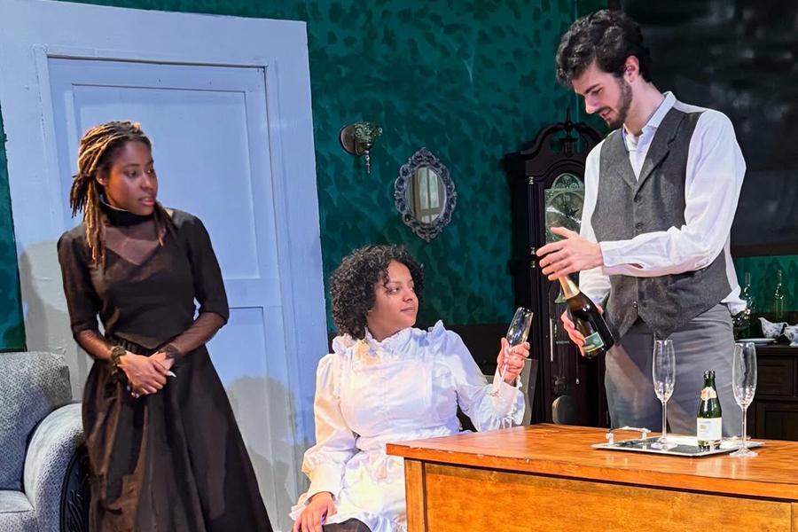 A man in a vest pours a bottle of wine for a seated woman in a white dress while another woman in black looks on in a scene from Henrik Ibsen's play "Ghosts."