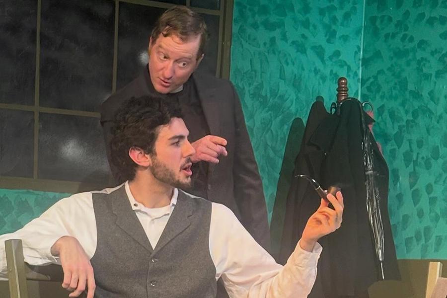 A man holding a smoking pipe looks away as a man dressed as a priest points at him with a critical expression in a scene from Henrik Ibsen's play "Ghosts."