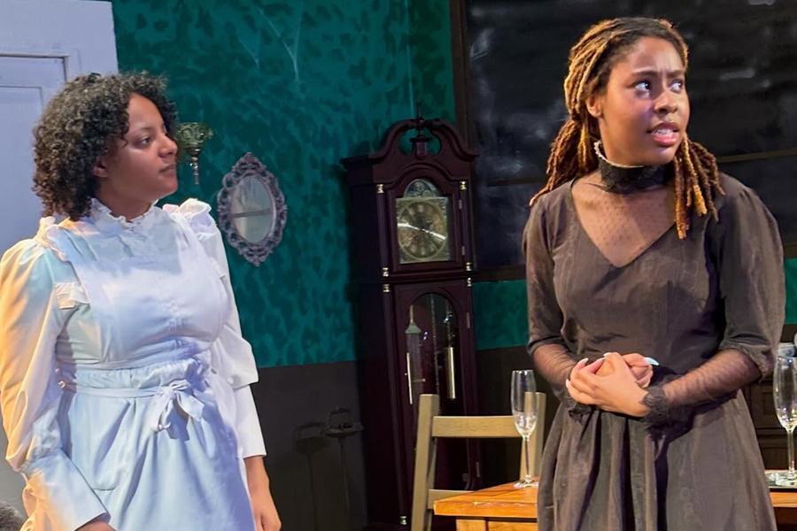 A woman in a white dress stands looking toward another woman who's wearing a dark dress and looks away with a troubled, tense expression in a scene from Henrik Ibsen's play "Ghosts."