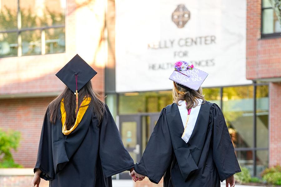 Two graduates hold hands in front of the Kelly Center.
