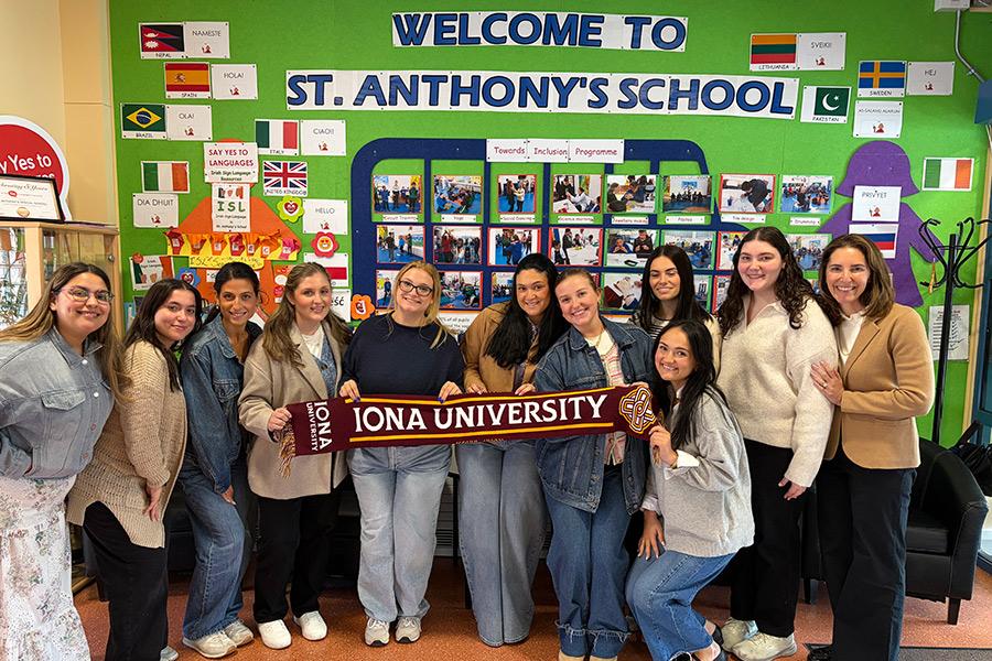 A group of Iona University students and faculty smile while holding a university scarf in front of a colorful "Welcome to St. Anthony's School" bulletin board.