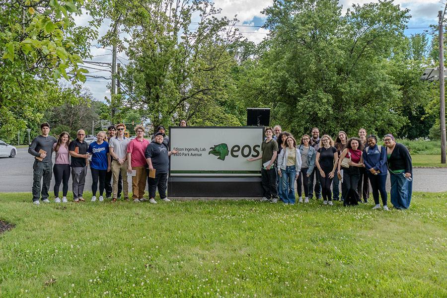 Iona University students and faculty smiling together while posing outdoors on either side of the Eos "Edison Ingenuity Lab" sign during their facility tour.