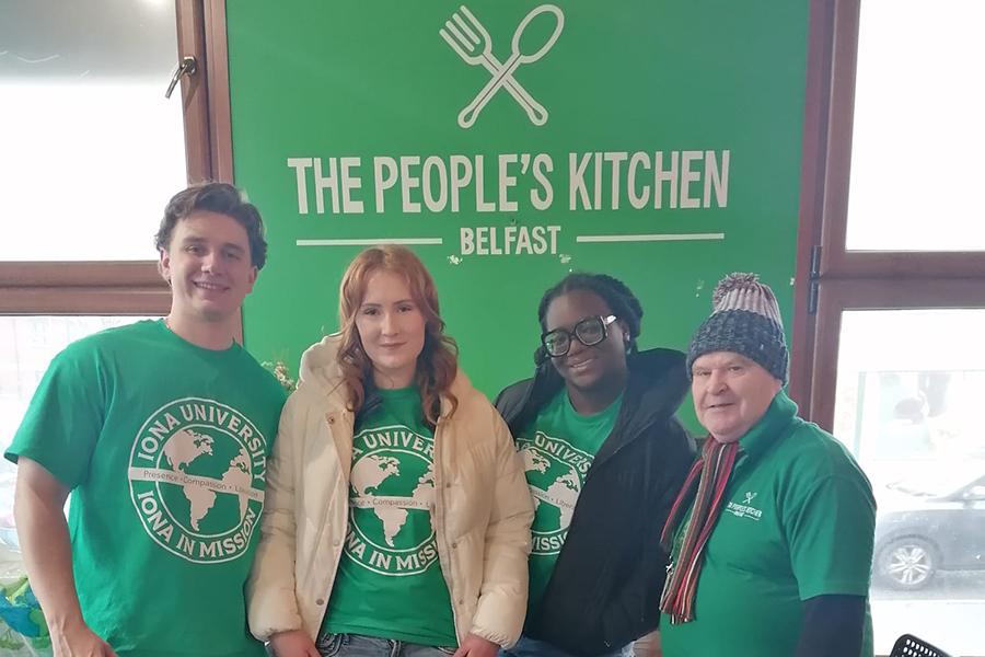 Four volunteers wearing Iona University in Mission shirts pose together in front of a green sign that reads "The People's Kitchen Belfast."