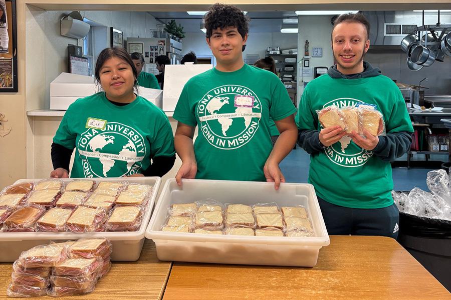 Three volunteers wearing Iona University in Mission shirts pose behind tables filled with freshly prepared sandwiches.