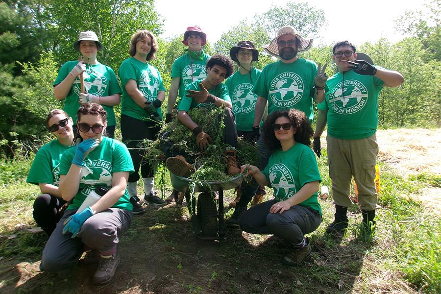 A group of students wearing matching Iona University in Mission green shirts pose outdoors around a wheelbarrow filled with grass brush.