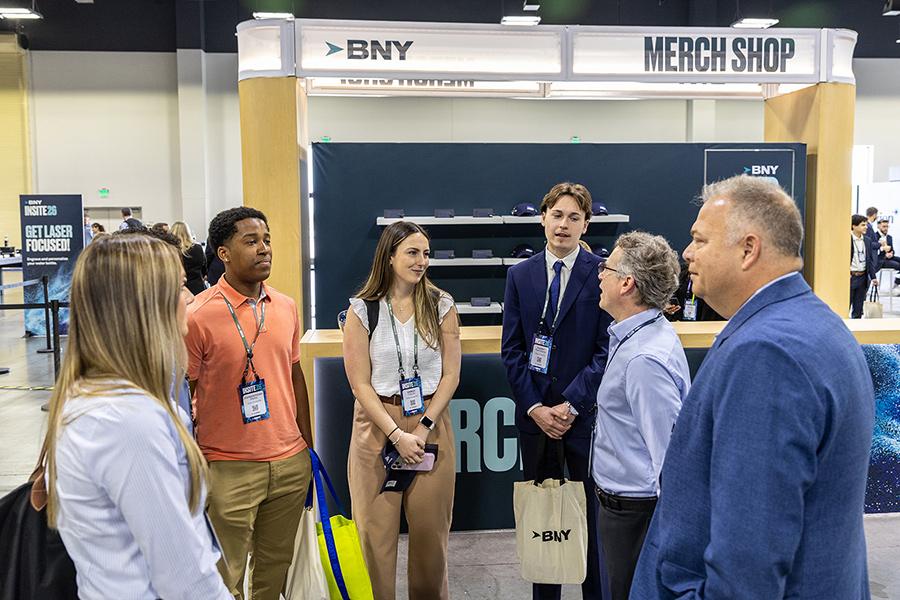 Iona University students converse with alumnus Christopher Fulco '89 and a conference leader near a BNY merch shop display.