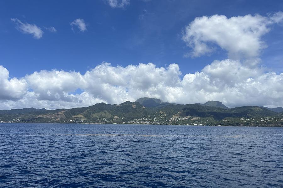 A scenic view from the water showing lush green mountains on the coastline of Dominica under a clear blue sky with white clouds.