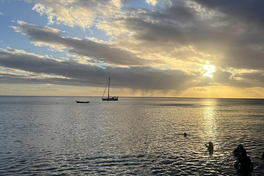 Dominica's Mero Beach at sunset with boats anchored on the water and people swimming in the foreground under a cloudy sky.