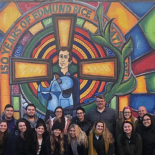 Students pose in front of a stained glass window of Edmund Rice that reads 150 years of Edmund Rice during a 2018-19 Winter trip.