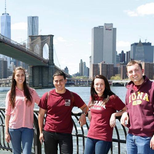 Four students in Iona gear stand in front of the river with NYC skyline and bridge in background.
