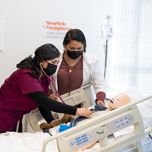 Two Iona nursing students practice skills on a human mannequin with NYP Iona School logo on the wall.