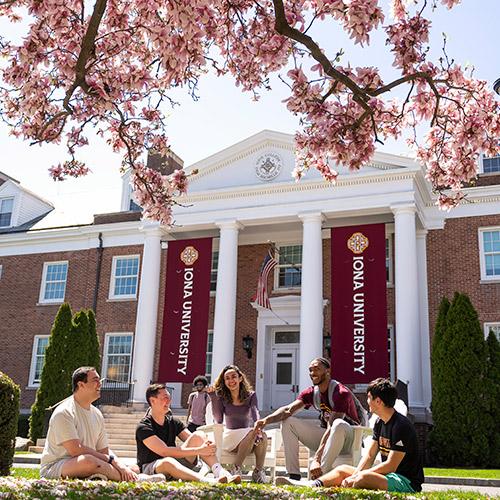 A group of friends sit in front of McSpedon Hall on a sunny day.