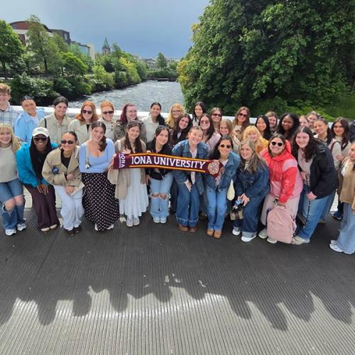 A large group of Iona University study abroad students and faculty pose together on a pedestrian bridge over a river in Ireland while holding up a university scarf.