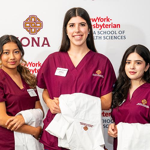Three nursing students with their white coats in front of an Iona step and repeat.