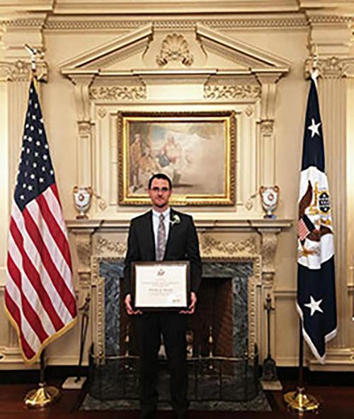 Nicholas Barnett posing with his Edward R. Murrow award, flanked with an American flag on his right and a Washington D.C. flag on his left.