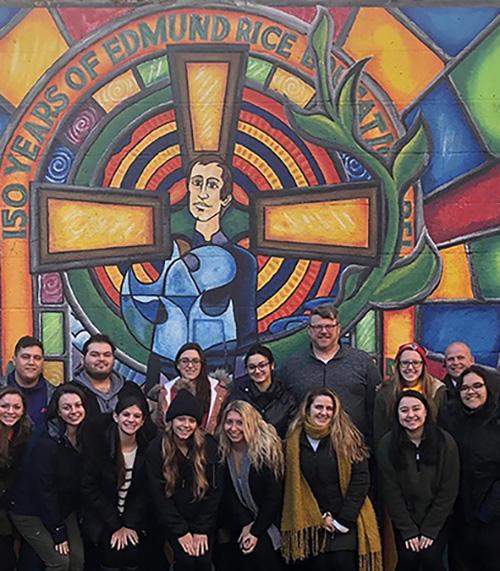 Students pose in front of a stained glass window of Edmund Rice that reads 150 years of Edmund Rice during a 2018-19 Winter trip.