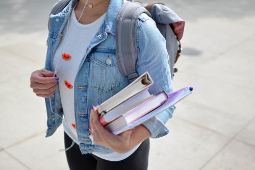 A girl in a jacket stands while carrying school books in her arm.