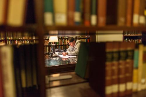 A student in a baseball cap studies in the library and we see him through an opening in the stacks of books.