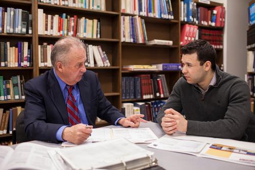 Two men sit together at a table discussing paperwork.