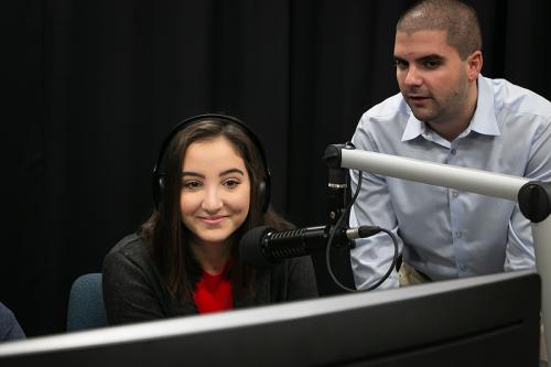 A student sits with headphones on in front of a microphone with a staff member standing on her right side.