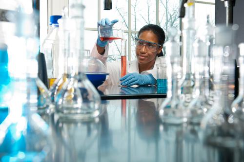 A student in a lab coat and goggles pours a liquid into a graduated cylinder.