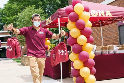 An orientation leader carries tote bags and swag near a table with balloons to welcome new Iona students.