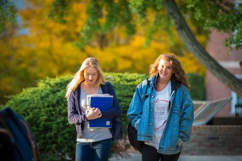 Two students walk on campus during early autumn in golden sunlight. One wears a jean jacket and the other carries books.