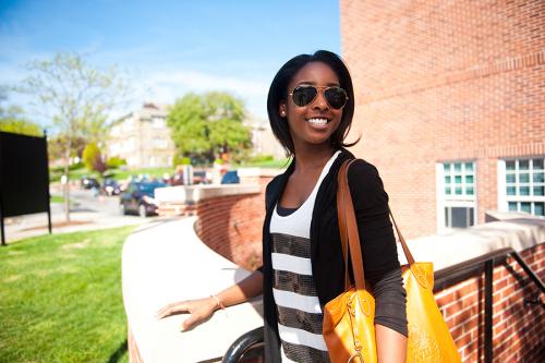 A student stands outside of the Starbucks on campus. She wears sunglasses and smiles.