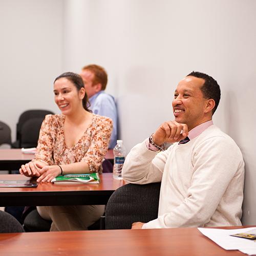 A student in a white sweater with a tie on leans on his hand and smiles during class.