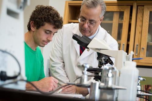 A professor in a lab coat helps a student at the microscope.