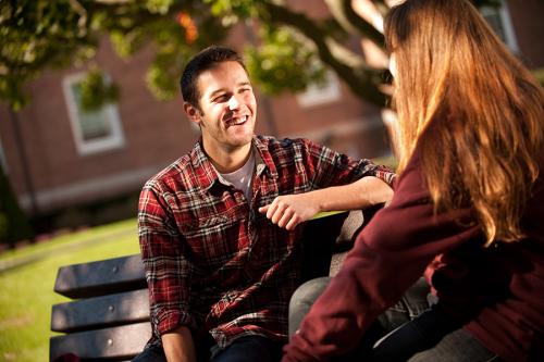 A student in a flannel shirt sits on a bench and talks with another student.
