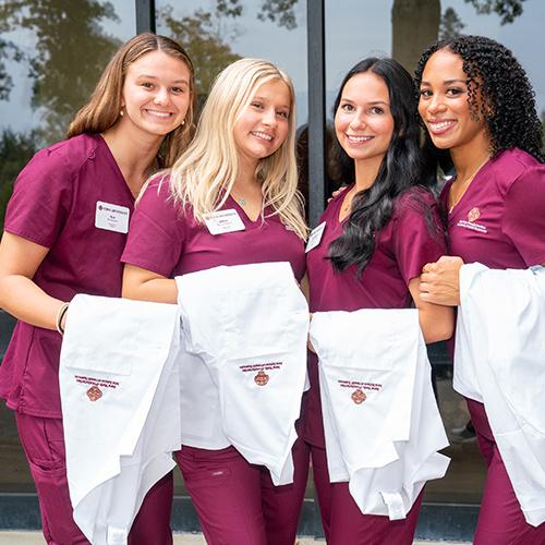 Four nursing students smile in their maroon scrubs and hold white coats.