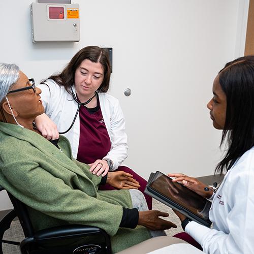 Two FNP students work with a mannequin patient in a wheelchair.