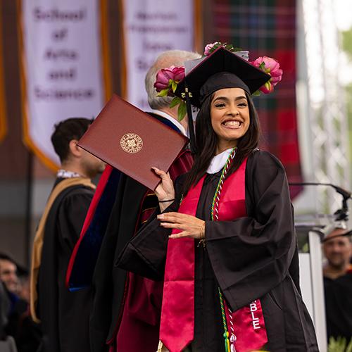 A student smiling and holding up their diploma at Commencement.